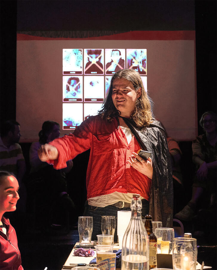 Lewie the game host, revealing the Traitors amongst the group. Lewie is in a black cape. Behind him is a scree. with the eliminated players.