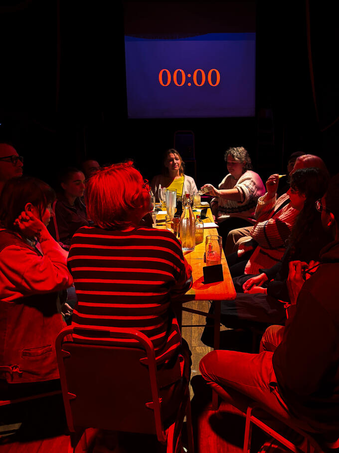 A group of people, having a roundtable discussion choosing who they thing the Traitor amongst the group is. Bathed in red light. A timer behind them has counted doen to zero.